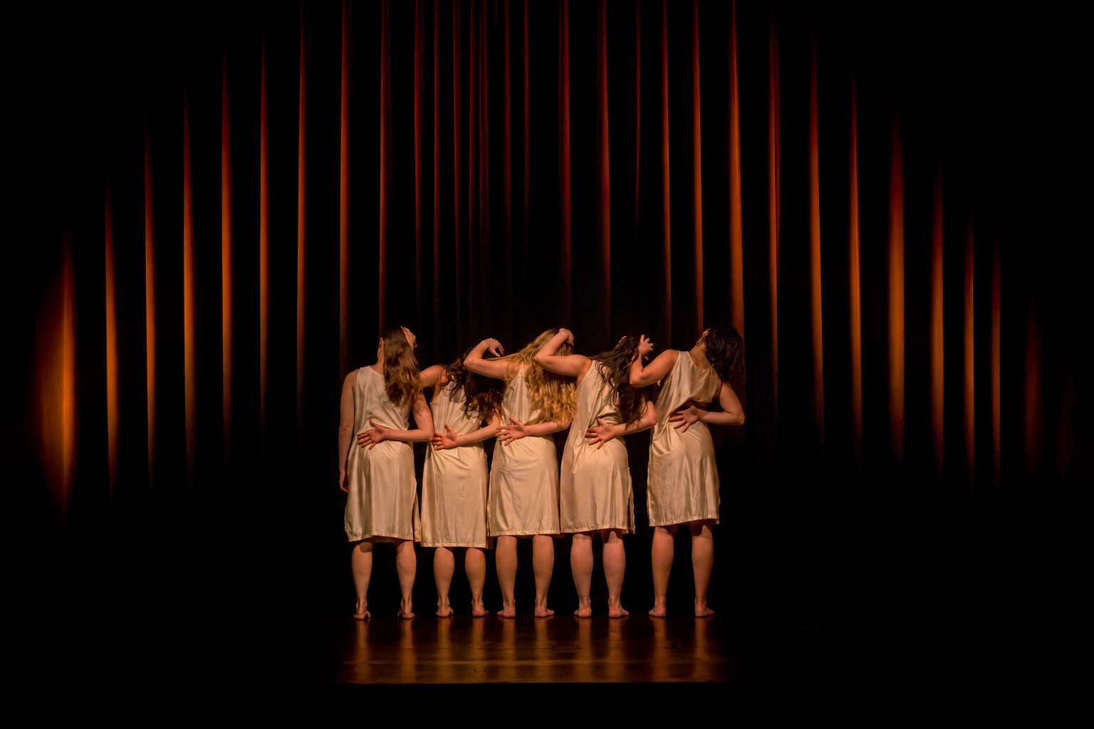 Six dancers seen from behind in beige dresses, arms intertwined, before a red curtain