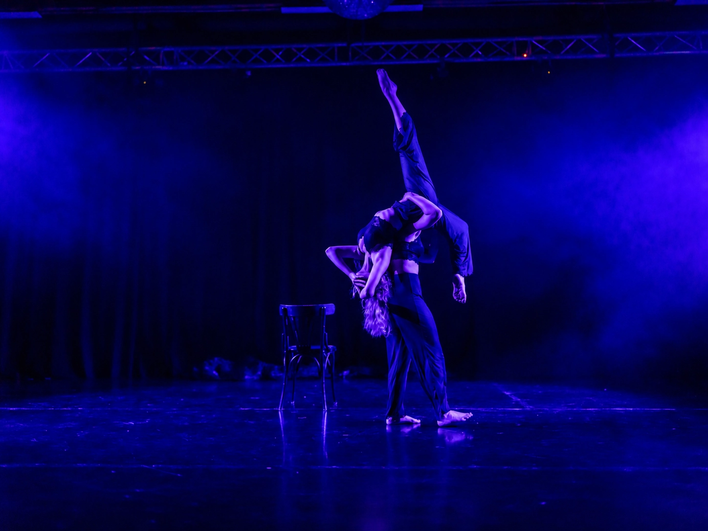 Duet under blue light, one dancer upside down with a leg pointing upward, a chair on stage