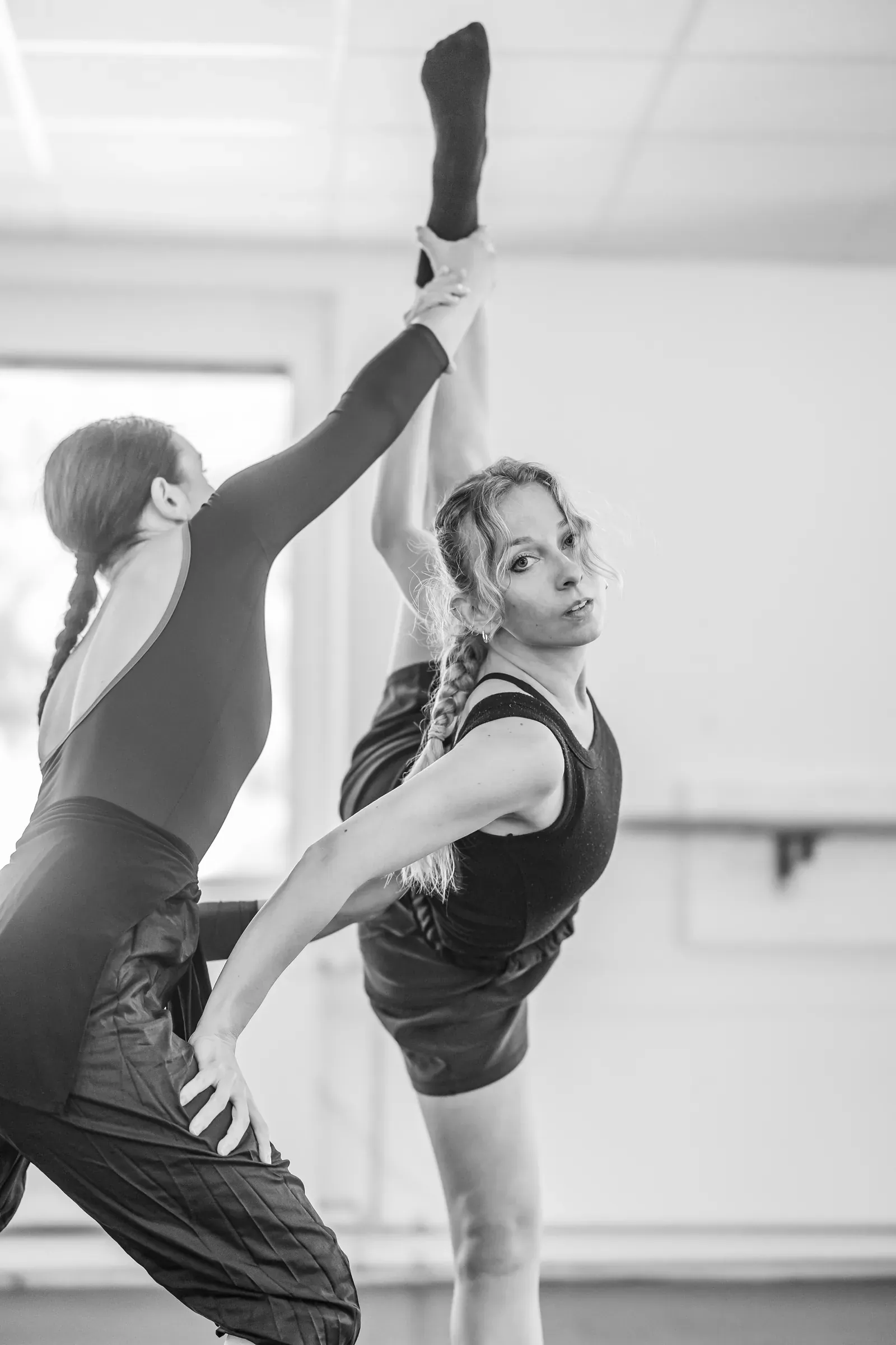 Lucie Marcellin stretching, her leg held vertically by a partner, in the studio (black and white)