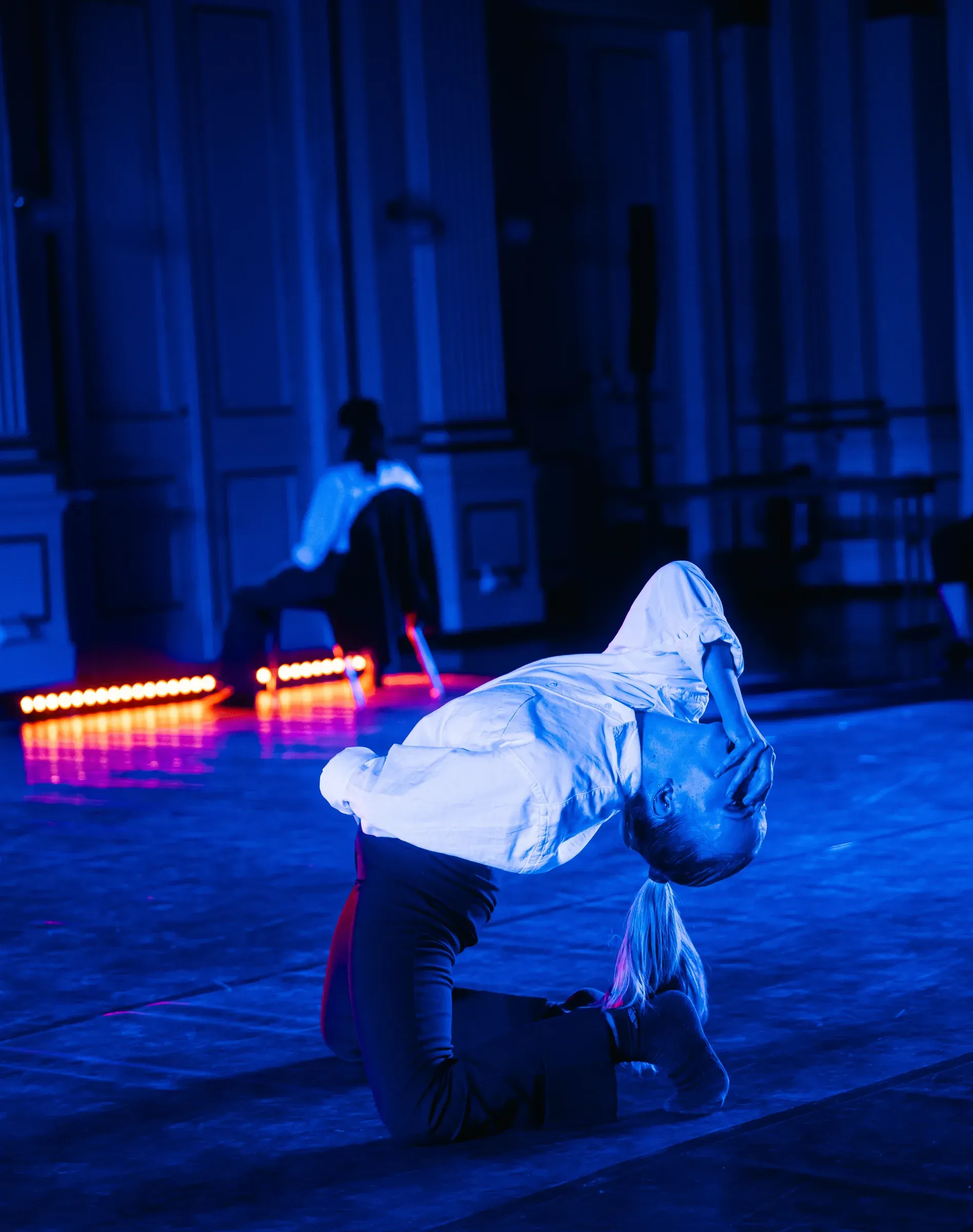 Dancer kneeling, back arched, in a white shirt under blue light